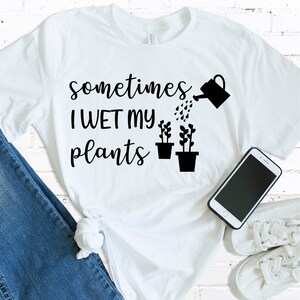 May include: White t-shirt with black text that reads "sometimes I WET my plants" and a black watering can pouring water on two black potted plants.
