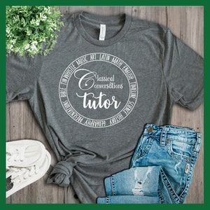 Puede incluir: Una camiseta gris con un dise&ntilde;o circular blanco que dice "Classical Conversations Tutor" y enumera materias como "Biblia, Tin Whistle, M&uacute;sica, Arte, Lat&iacute;n, Matem&aacute;ticas, Ingl&eacute;s, Cronolog&iacute;a, Ciencias, Historia, Geograf&iacute;a, Presentaciones".
