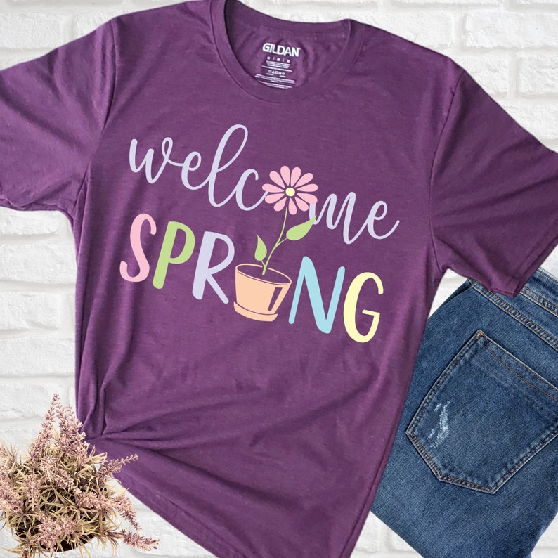 K&ouml;nnte beinhalten: Lilafarbenes T-Shirt mit dem Text "welcome spring" in bunten Buchstaben. Eine Blume in einem Topf ist Teil des Textdesigns.