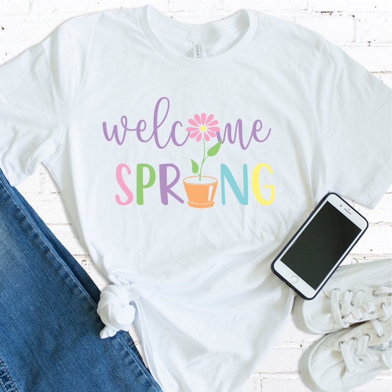 K&ouml;nnte beinhalten: Wei&szlig;es T-Shirt mit dem Text "welcome spring" in bunten Buchstaben. Eine rosa Blume mit gr&uuml;nem Stiel w&auml;chst aus einem braunen Topf.