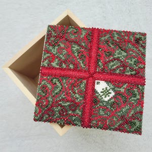Può includere: Una scatola quadrata di legno con un coperchio decorato con perline. Il coperchio presenta un motivo rosso e verde, un motivo a nastro rosso e un piccolo emblema bianco e verde. La scatola è aperta, rivelando l'interno in legno naturale.