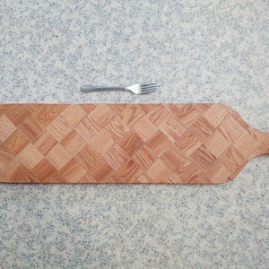 May include: A long, wooden serving board with a woven pattern. The board has a handle on one end and is made of light brown wood.