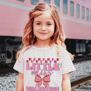Puede incluir: Una camiseta rosa claro con el texto "Little Miss Valentine" en rosa y rojo. La camiseta presenta un lazo y un patrón de cuadros. Una niña pequeña lleva la camiseta delante de un tren rosa.