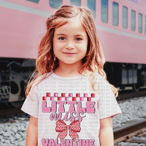 Puede incluir: Una camiseta rosa claro con el texto "Little Miss Valentine" en rosa y rojo. La camiseta presenta un lazo gráfico y un patrón de cuadros. El fondo incluye un tren rosa y vías de ferrocarril.