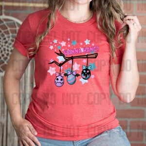Puede incluir: Camiseta roja con un gráfico de una rama con tres adornos colgantes. Los adornos son de color rosa, azul y morado con un diseño de calavera y huesos cruzados. El texto "Wanna Hang?" está escrito en la rama.