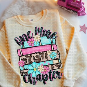 May include: Cream-colored sweatshirt with the text "One More Chapter" in black script. The design features a stack of books with floral and leopard print accents. A pink toy train and star-shaped decorations are in the background.