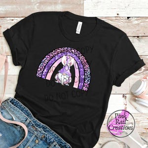 Pode incluir: Camiseta preta com um arco-íris roxo, rosa e branco com um gnomo usando um chapéu roxo e orelhas de coelho brancas em pé no centro. O texto "Do Not Copy" está abaixo do arco-íris.