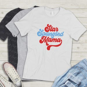 Puede incluir: Una camiseta blanca con el texto "Star Spangled Mama" en rojo y azul. La camiseta se muestra con otras camisetas en gris y negro, un par de vaqueros azules y zapatillas blancas.