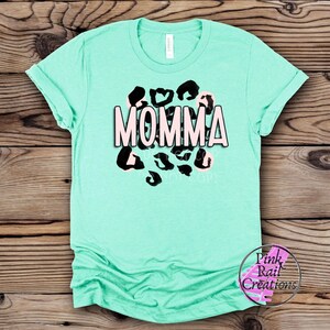 Pode incluir: Uma camiseta turquesa com um design de estampa de leopardo em preto e branco e a palavra "MOMMA" em letras rosa.