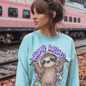 Puede incluir: Sudadera azul claro con un diseño de perezoso de dibujos animados y el texto "WOMP WOMP". El perezoso está rodeado por un círculo morado con un patrón de cuadros y estrellas coloridas. Un tren rosa y vías de ferrocarril se ven al fondo.