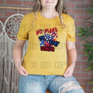 May include: Mustard yellow t-shirt with the phrase "NO PLANS TO STAY SOBER" in red, white, and blue. The graphic includes a cocktail with a straw and a small umbrella. The model is wearing blue jeans.