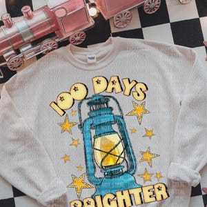 May include: Cream sweatshirt featuring the text "100 Days Brighter" and a lantern graphic. The design includes yellow stars and a blue lantern. A toy train set is visible in the background.