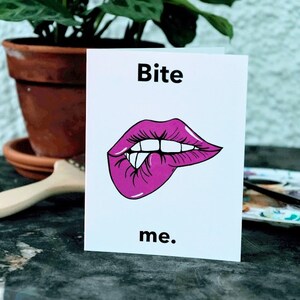 May include: A white card with the text "Bite me." in black lettering. A pink cartoon mouth with teeth is biting its lower lip.