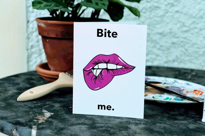 May include: A white card with the text "Bite me." in black lettering. A pink cartoon mouth with teeth is biting its lower lip.