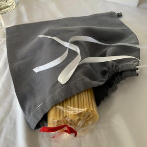 May include: A grey fabric drawstring bag with white ribbon ties, partially open to reveal a clear plastic package of long, thin, yellow dried pasta noodles inside. The pasta package features a small red ribbon tied at the bottom end.