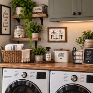 May include: A laundry room setup featuring two white LG washing machines, a wooden countertop, and wall-mounted shelves. Decor includes signs reading A clean home is a happy home, what the FLUFF, and WASH DRY FOLD REPEAT, plus baskets, plants, and containers.