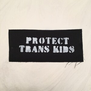 May include: A black rectangular patch with the words "PROTECT TRANS KIDS" in white, block letters. The patch is set against a cream-colored background.
