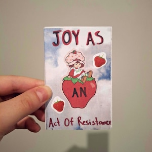 May include: A rectangular sticker with a sky blue background and the words "JOY AS AN Act Of Resistance" in red and black. The sticker features a cartoon strawberry character sitting on a large red strawberry.