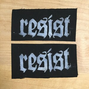 May include: Two black fabric patches with the word "resist" written in white, stylized lettering.