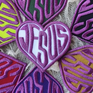 Peut inclure: Sept patchs en forme de cœur violets avec le mot "JESUS" écrit dans différents styles et couleurs. Les patchs sont disposés en cercle sur un fond de tissu blanc.