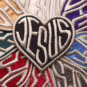 May include: A colorful arrangement of embroidered patches in the shape of hearts, each with the word "JESUS" spelled out in a different color and style. The patches are arranged in a circular pattern, with the largest patch in the center.
