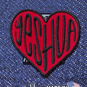 Puede incluir: Un parche rojo en forma de corazón con texto bordado en negro que dice "JESUS" en una fuente estilizada. El parche está sobre un fondo de mezclilla azul. El texto "ONLYGOD OUTFITTERS" y "PATCH" son visibles sobre el corazón. El texto "DIY SEW OR IRON-ON" también es visible sobre el corazón. El texto "Thank You MADE IN WASHINGTON" es visible debajo del corazón.