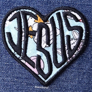 Puede incluir: Un parche en forma de corazón con la palabra "JESUS" bordada en negro sobre un fondo floral azul y blanco.