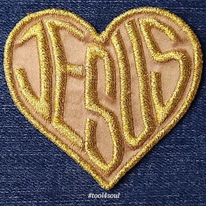 May include: A heart-shaped patch with the word "JESUS" embroidered in gold thread on a beige fabric background. The patch is on a dark blue denim background.