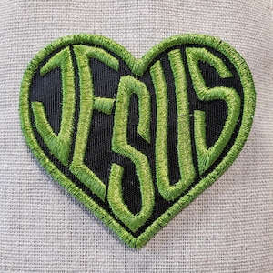 May include: Heart-shaped patch with the word "JESUS" embroidered in bright green thread on a black background. The patch has a green border and is set against a textured, light-colored fabric.