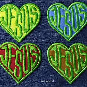 May include: Four heart-shaped patches with the word "JESUS" written in green letters on a yellow, blue, red, and brown background. The patches are on a blue denim background. #tool4soul