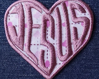 May include: Pink heart-shaped patch with the word "JESUS" embroidered in white on a pink and white checkered background.