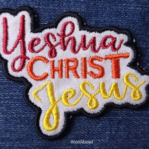 Peut inclure: Écusson brodé avec les mots "Yeshua CHRIST JESUS" en écriture rouge, orange et jaune. L'écusson a un fond blanc avec une bordure noire, sur un fond en denim bleu. Le texte "#tool4soul" est en bas.