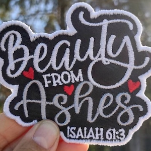 May include: Embroidered patch with the words "Beauty From Ashes" in white script on a black background. Features three small red hearts and the Bible verse Isaiah 61:3. The patch has a white border.