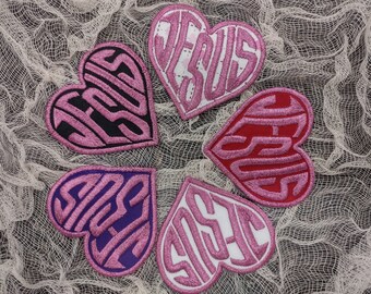 Op de afbeelding: Vijf roze hartvormige patches met het woord "Jesus" geschreven in een gestileerd lettertype. De patches zijn gemaakt van stof en hebben een roze en witte kleurstelling.