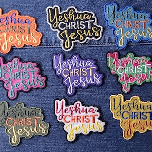 May include: Nine embroidered patches featuring the text Yeshua CHRIST Jesus in various color combinations, including orange, black, blue, pink, purple, green, olive, white, and gold thread, arranged in a grid on a blue denim fabric background.