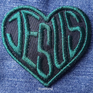 Peut inclure: Un patch brodé en forme de cœur vert avec le mot "JESUS" sur un tissu bleu foncé.