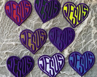 Op de afbeelding: Acht hartvormige patches met het woord "JESUS" erop geschreven in verschillende kleuren en stijlen. De patches zijn gemaakt van stof en hebben een kleurenschema in paars, rood, groen, geel, blauw en wit.