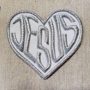 May include: A white heart-shaped patch with the word "JESUS" embroidered in silver thread.