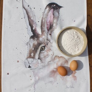 Könnte beinhalten: Ein weißes Geschirrtuch mit einer Aquarell-Illustration eines braunen und weißen Hochlandhasen. Der Hase schaut nach links. Das Geschirrtuch hat die Worte "Highland Hare" in Grau unten aufgedruckt. Auf dem Geschirrtuch liegen zwei braune Eier und eine Schüssel mit weißem Mehl.