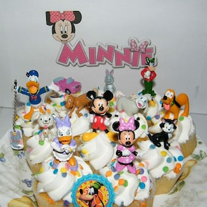 Puede incluir: Un plato de cupcakes decorados con chispas de colores y varias figuras de personajes de Disney, incluyendo Mickey Mouse, Minnie Mouse, Donald Duck, Goofy y Pluto. Los cupcakes están cubiertos con glaseado blanco y el plato tiene un diseño floral.