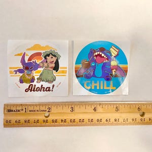 Może przedstawiać: Dwa okrągłe naklejki z Stitch'em z Lilo & Stitch. Pierwsza naklejka jest niebieska z Stitch'em w kwiecistym naszyjniku i napisem "Chill". Druga naklejka jest pomarańczowa i ż&oacute;łta z Stitch'em i dziewczyną w kwiecistych naszyjnikach i napisem "Aloha!"