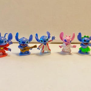 May include: Five blue and pink Stitch figurines. The Stitch figurines are wearing different outfits, including a red shirt, a blue shirt with a guitar, a striped shirt, a pink and white dress, and a green grass skirt.