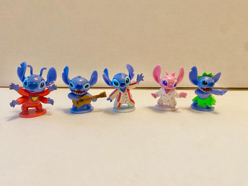 May include: Five blue and pink Stitch figurines. The Stitch figurines are wearing different outfits, including a red shirt, a blue shirt with a guitar, a striped shirt, a pink and white dress, and a green grass skirt.