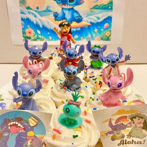 May include: A collection of blue and pink Stitch character cupcakes with sprinkles. The cupcakes are decorated with various Stitch figurines, including a surfing Stitch, a Stitch with a lei, and a Stitch with a pineapple. The cupcakes are on a white plate with a blue and white background.