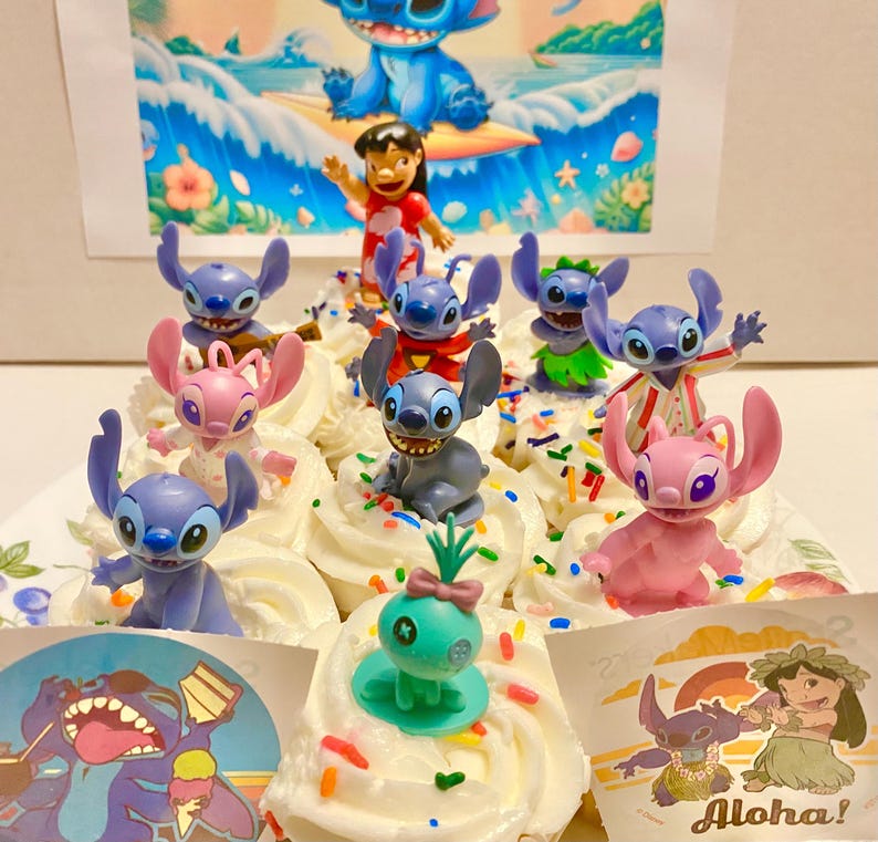 May include: A collection of blue and pink Stitch character cupcakes with sprinkles. The cupcakes are decorated with various Stitch figurines, including a surfing Stitch, a Stitch with a lei, and a Stitch with a pineapple. The cupcakes are on a white plate with a blue and white background.