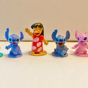 Może przedstawiać: Pięć małych plastikowych figurek postaci z filmu Disneya Lilo & Stitch. Postacie to Stitch, Angel, Lilo i dwie inne wersje Stitcha. Figurki stoją na białym tle.