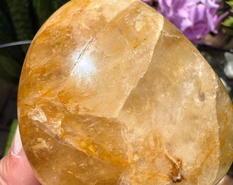 May include: A polished, rounded golden citrine gemstone with a translucent, crystalline structure. The stone features internal fractures, natural inclusions, and a warm, honey-yellow hue that reflects light across its smooth, curved surface.