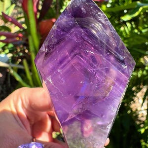 May include: A large, faceted amethyst crystal with a diamond-like shape, held in a hand. The crystal is a deep purple color with visible internal patterns. A matching amethyst ring is visible in the foreground, set in a gold band.
