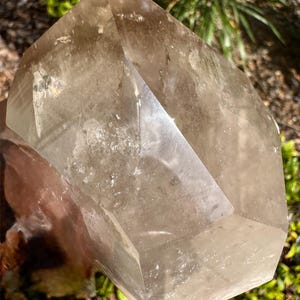 May include: A large, faceted smoky quartz crystal with a light brown hue. The crystal has a geometric shape with multiple flat surfaces and sharp edges. The crystal is displayed outdoors, with green foliage in the background.