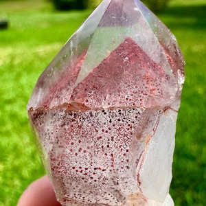 May include: A clear, pointed quartz crystal featuring a distinct internal phantom formation. The interior displays a concentrated layer of reddish-brown, speckled mineral inclusions that form a triangular shape within the transparent structure.